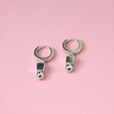 A pair of small hoop earrings with geometric charms attached, displayed against a pink background.