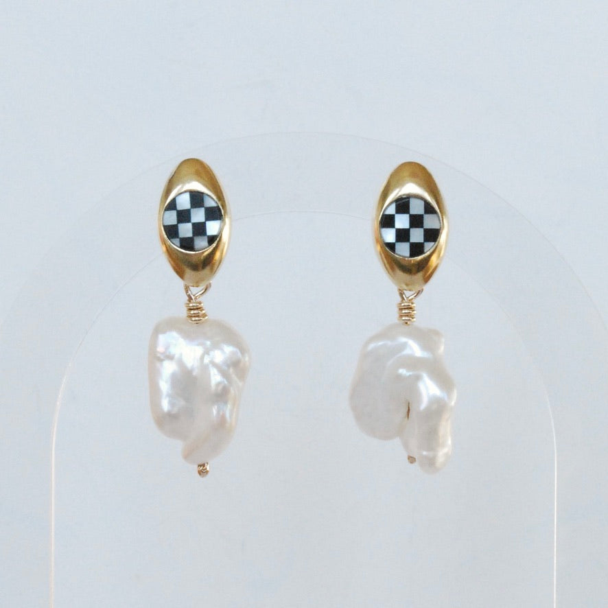 A pair of oval inlay pearl posts with baroque pearl dangles, with the earrings displayed in a gold-colored oval post and a checkered pattern on the inlay.