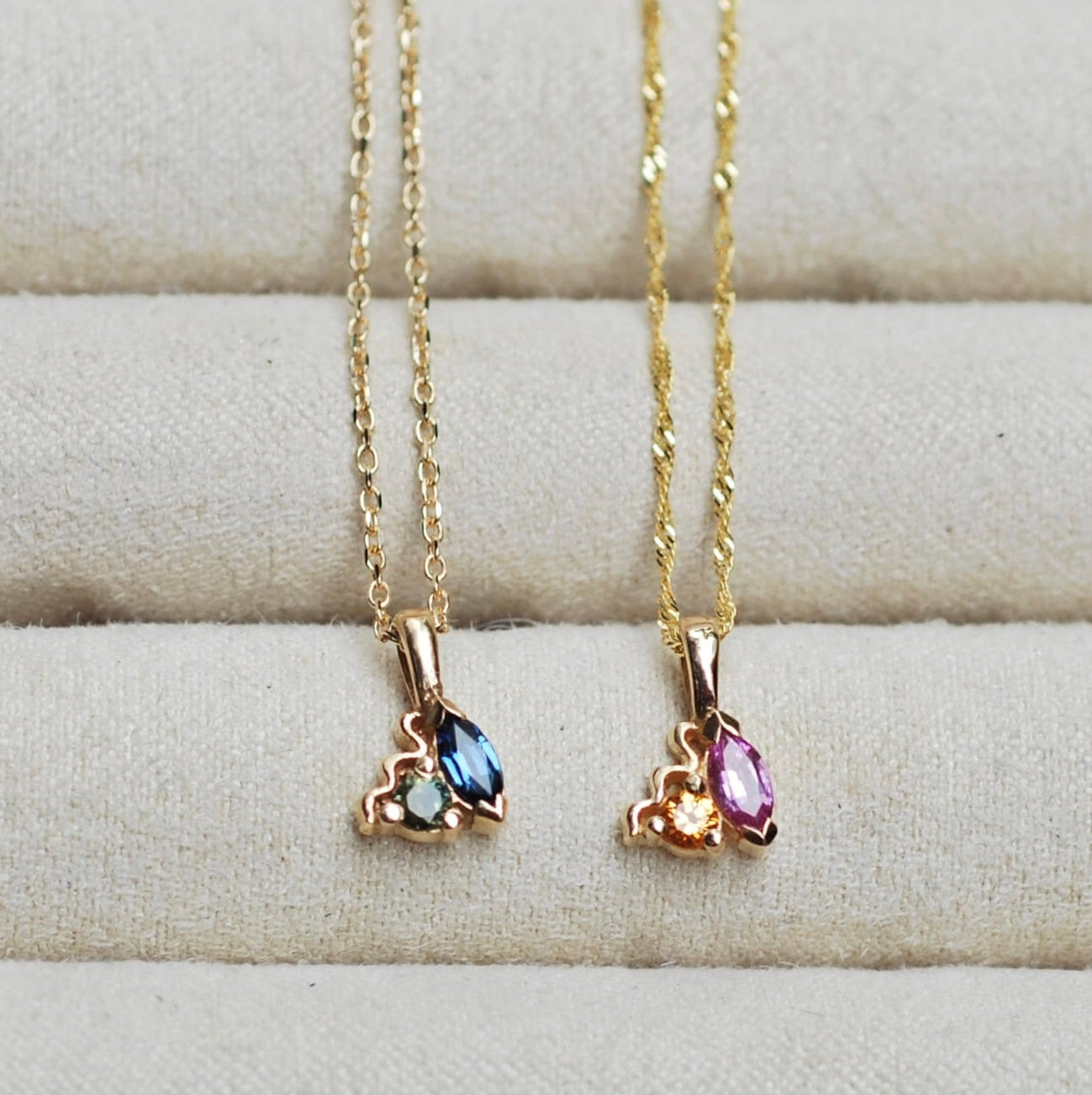 Two gold necklaces with sapphire gemstone pendants on a textured surface