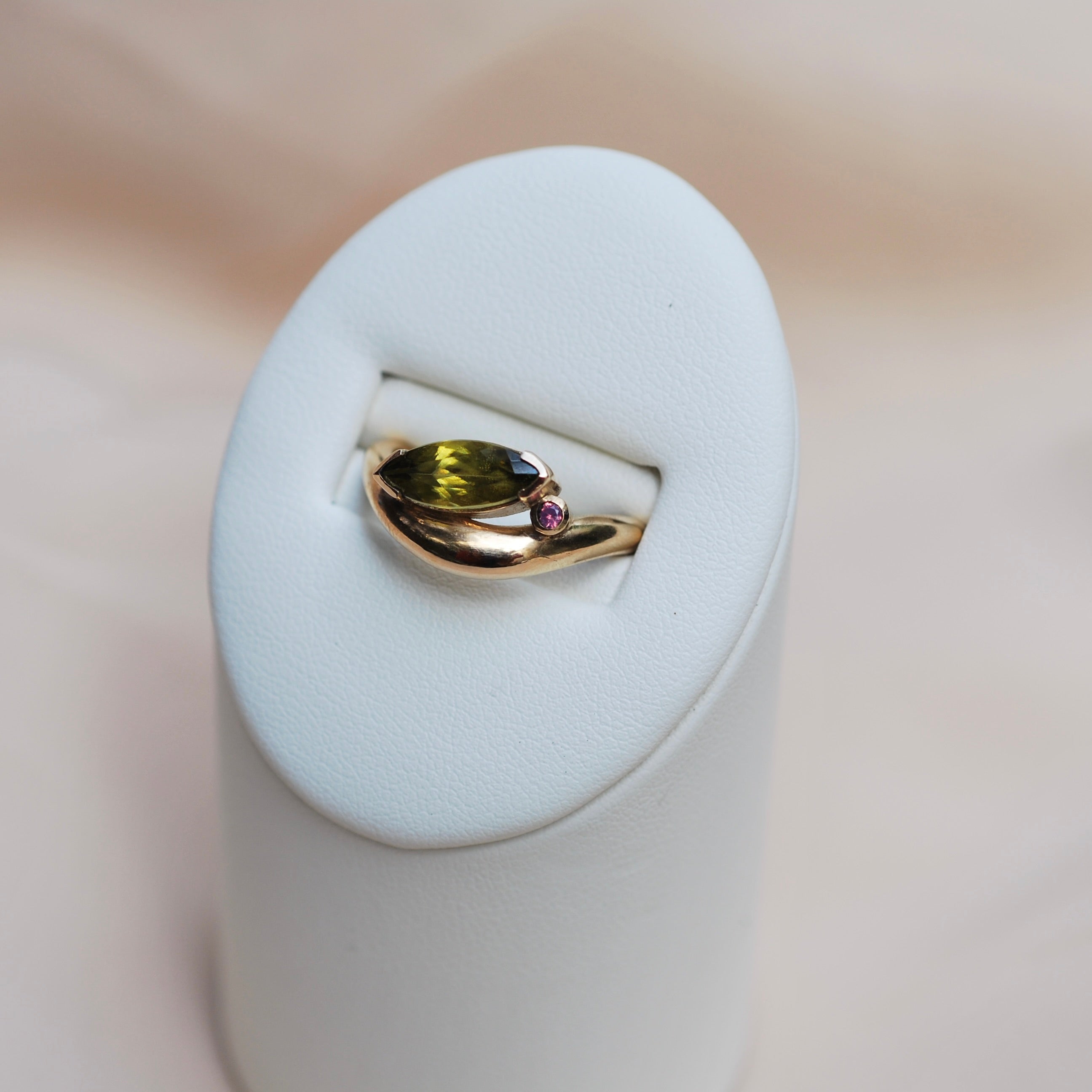 Gold ring with an organic band and a tilted green marquis stone sitting on a pink sapphire sitting in a white ring display