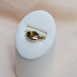 Gold ring with an organic band and a tilted green marquis stone sitting on a pink sapphire sitting in a white ring display