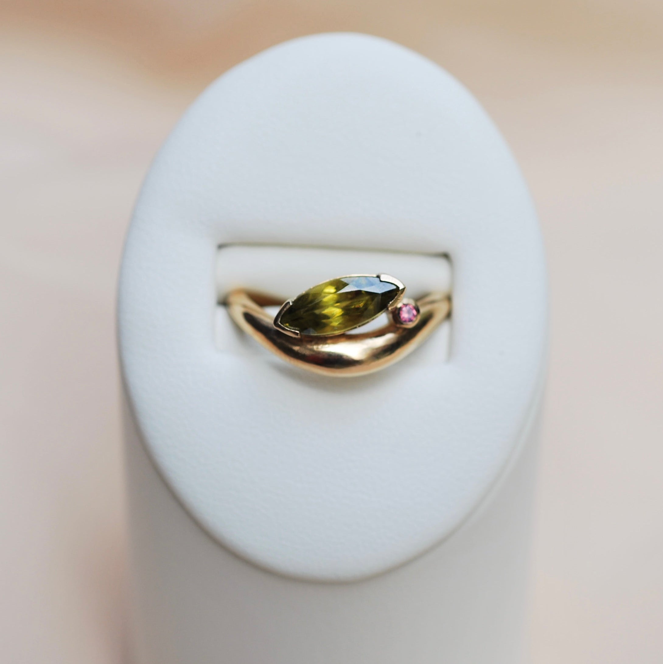 Gold ring with green sphene marquis and pink round sapphire on a white stand against a beige background