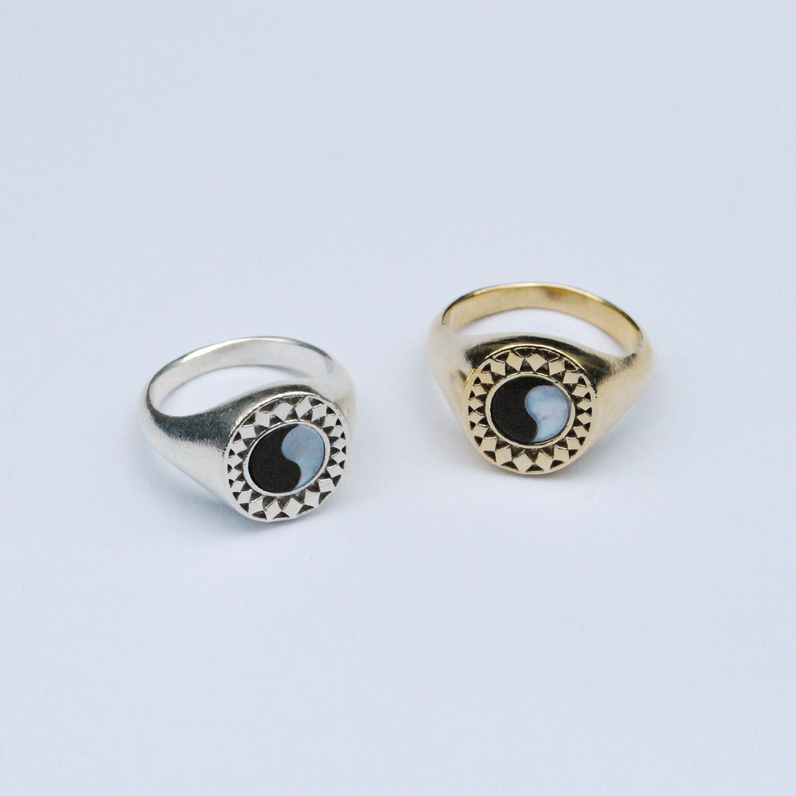Two rings on a grey background, one silver and one bronze, both with a black and white ying yang inlay stone surrounded by a star burst pattern