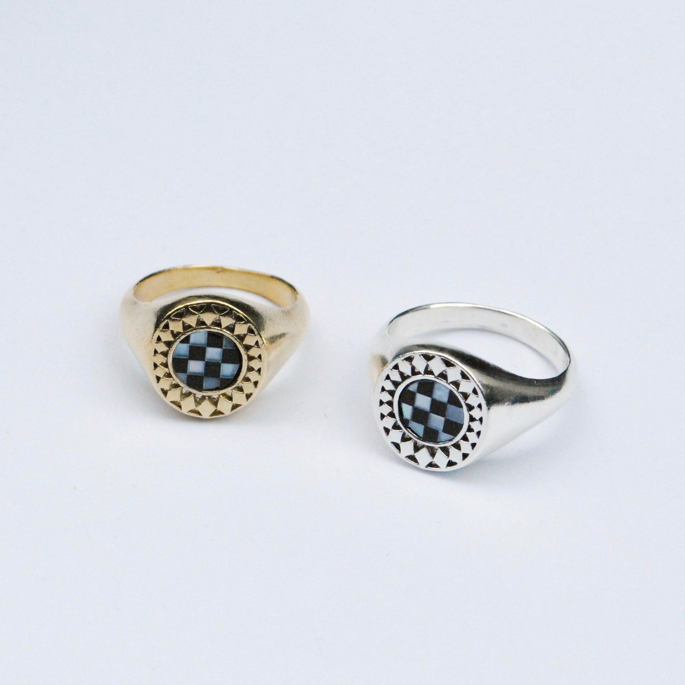 Two rings on a grey background, one silver and one bronze, both with a black and white checkerboard inlay stone surrounded by a star burst pattern