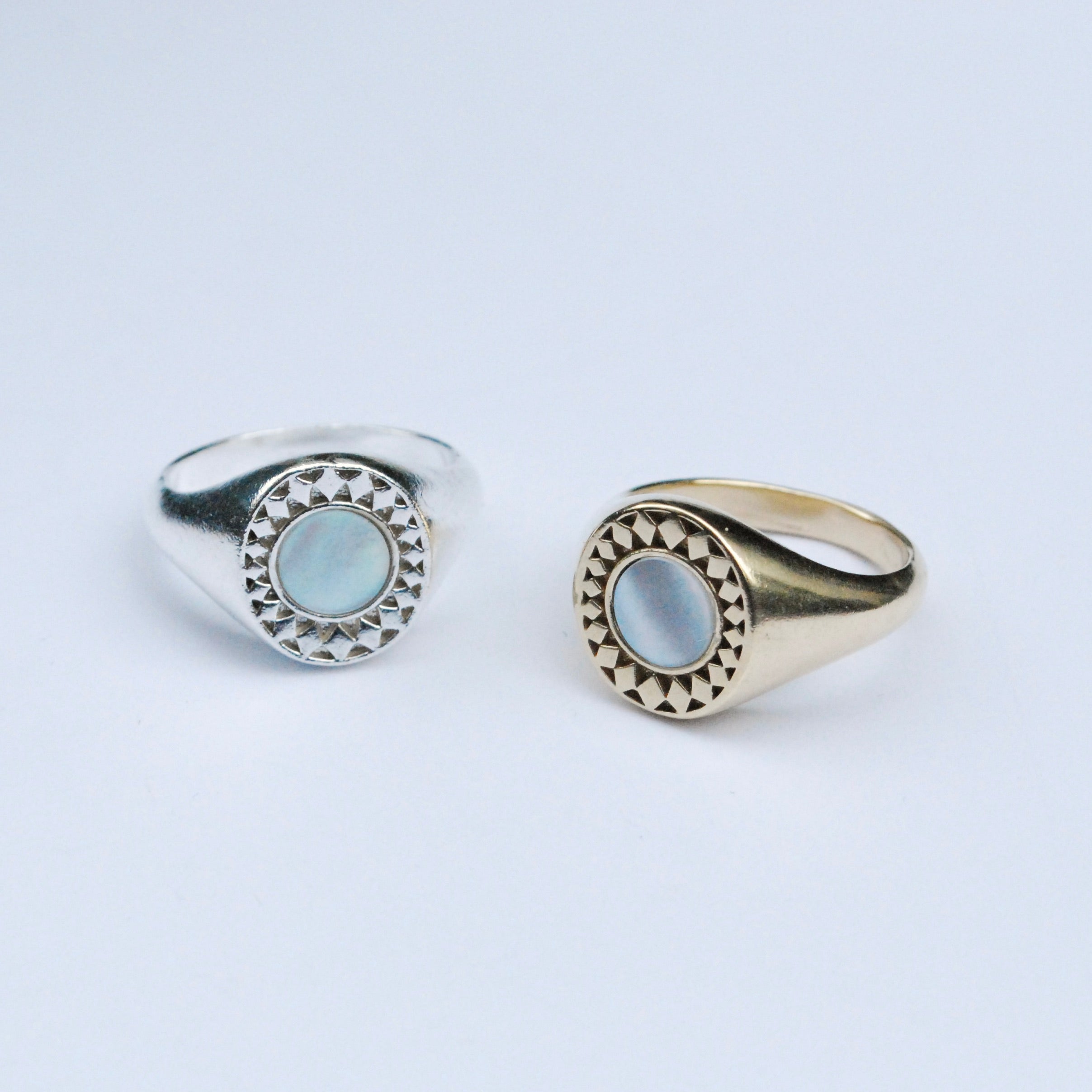 Two rings on a grey background, one silver and one bronze, both with a white mother of pearl inlay stone surrounded by a star burst pattern