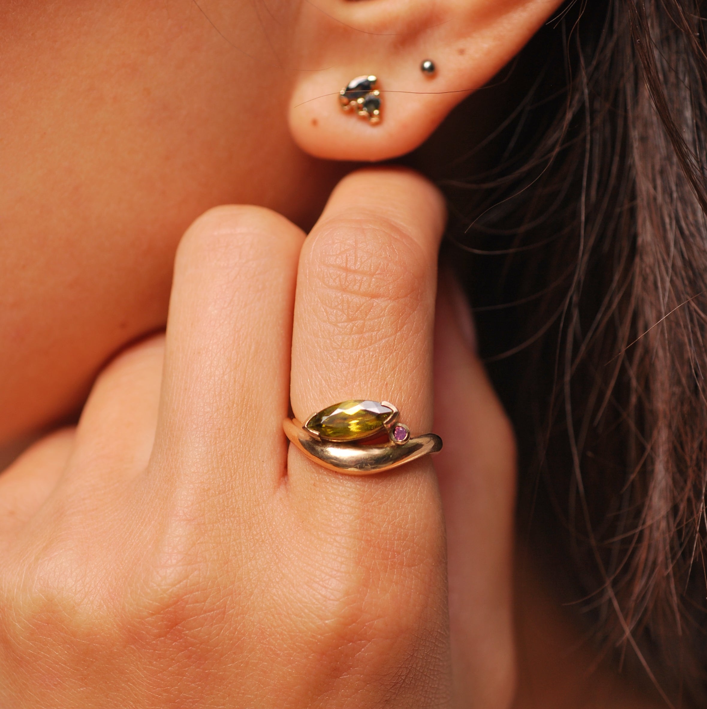 Close up of a hand wearing a gold ring with an organic swooped band and a tilted green marquis stone and pink accent stone