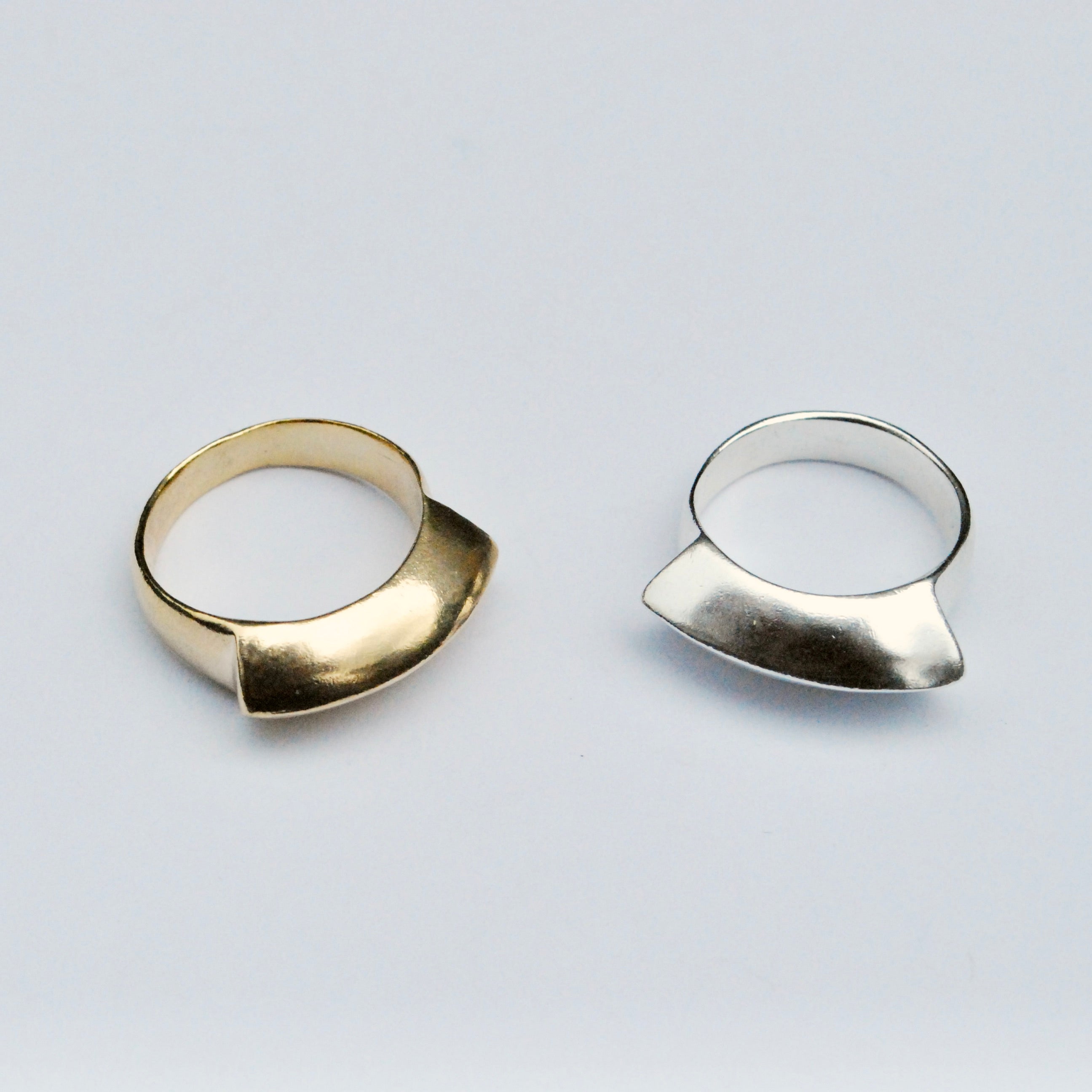 Two rings, one gold toned bronze and the other in silver, with a smooth, geometric design, displayed against a white background.