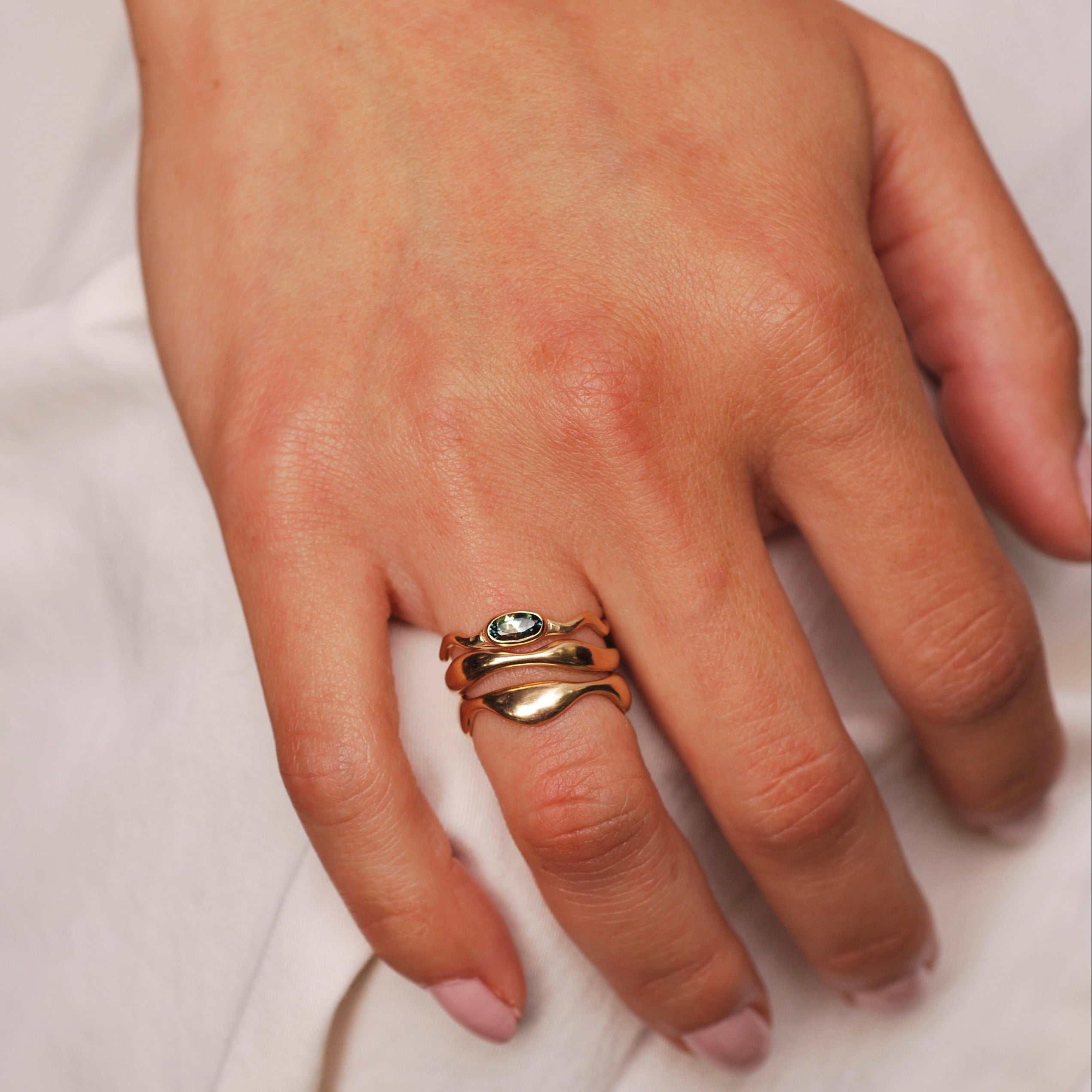 Hand wearing a stack of three organic wavy gold rings with a blue sapphire in one, on a white background