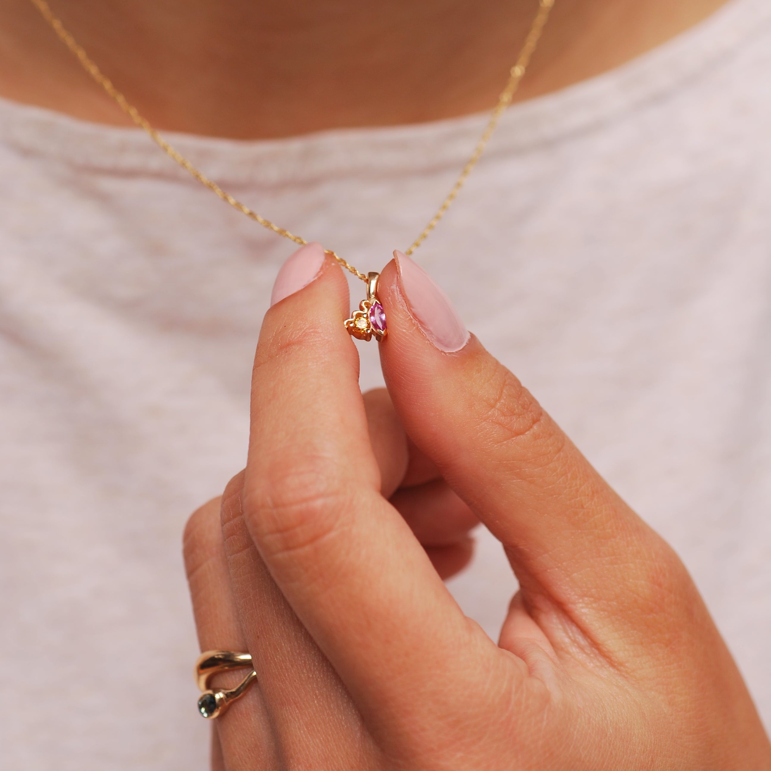 Hand holding a gold necklace with a pink and orange sapphire pendant against a neutral background