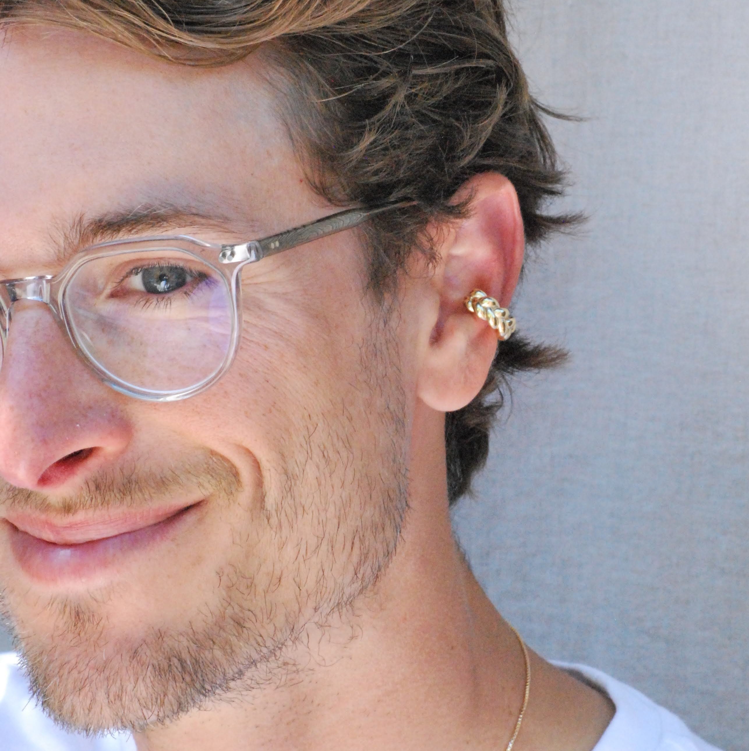 Close-up of a person wearing glasses and a braided gold ear cuff with a neutral background