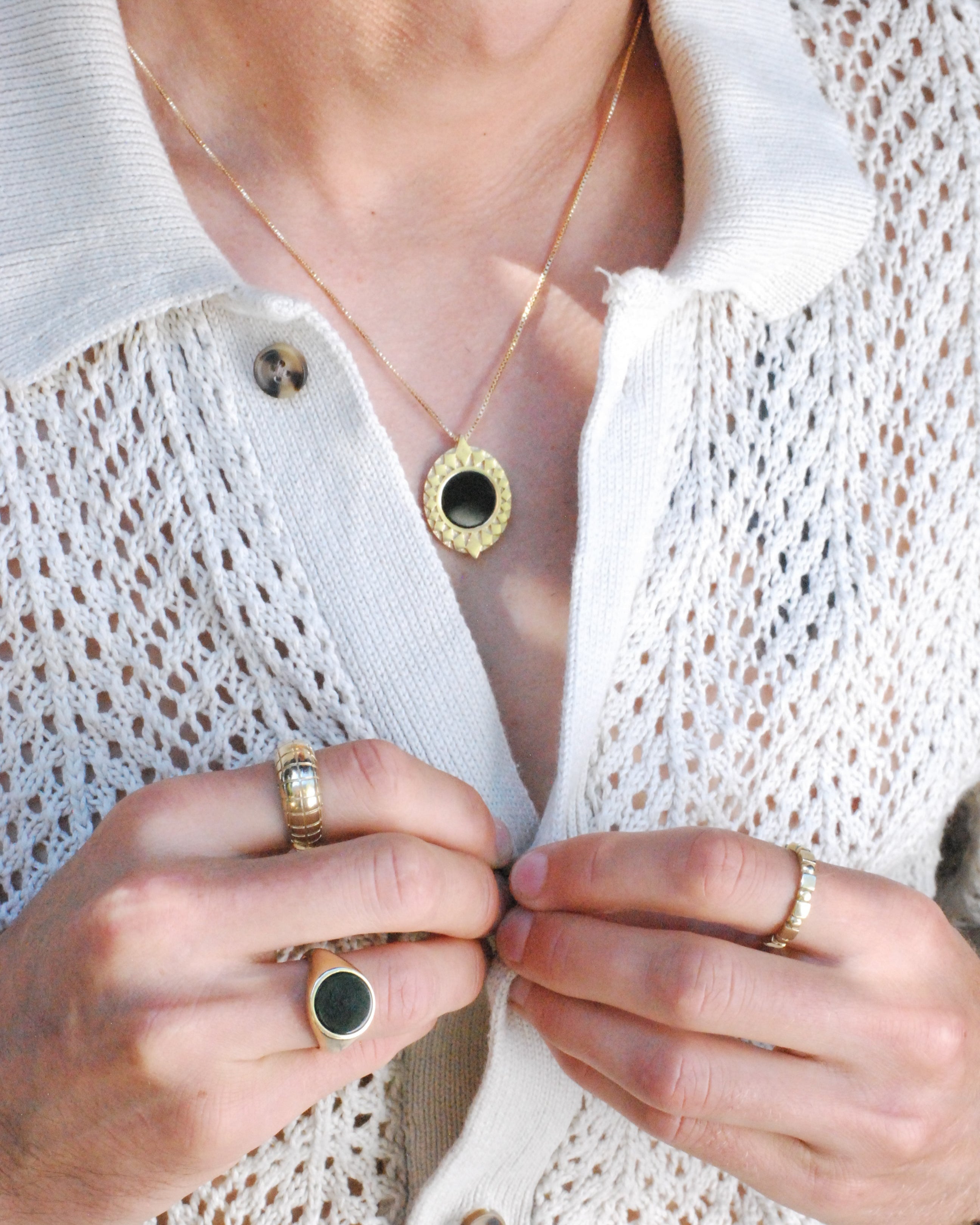 Close-up of a person wearing gold rings and a gold pendant with black stone on a white textured fabric background.