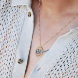Close-up of a gold necklace with a checkerboard pendant and starburst border on a person wearing a white woven top.
