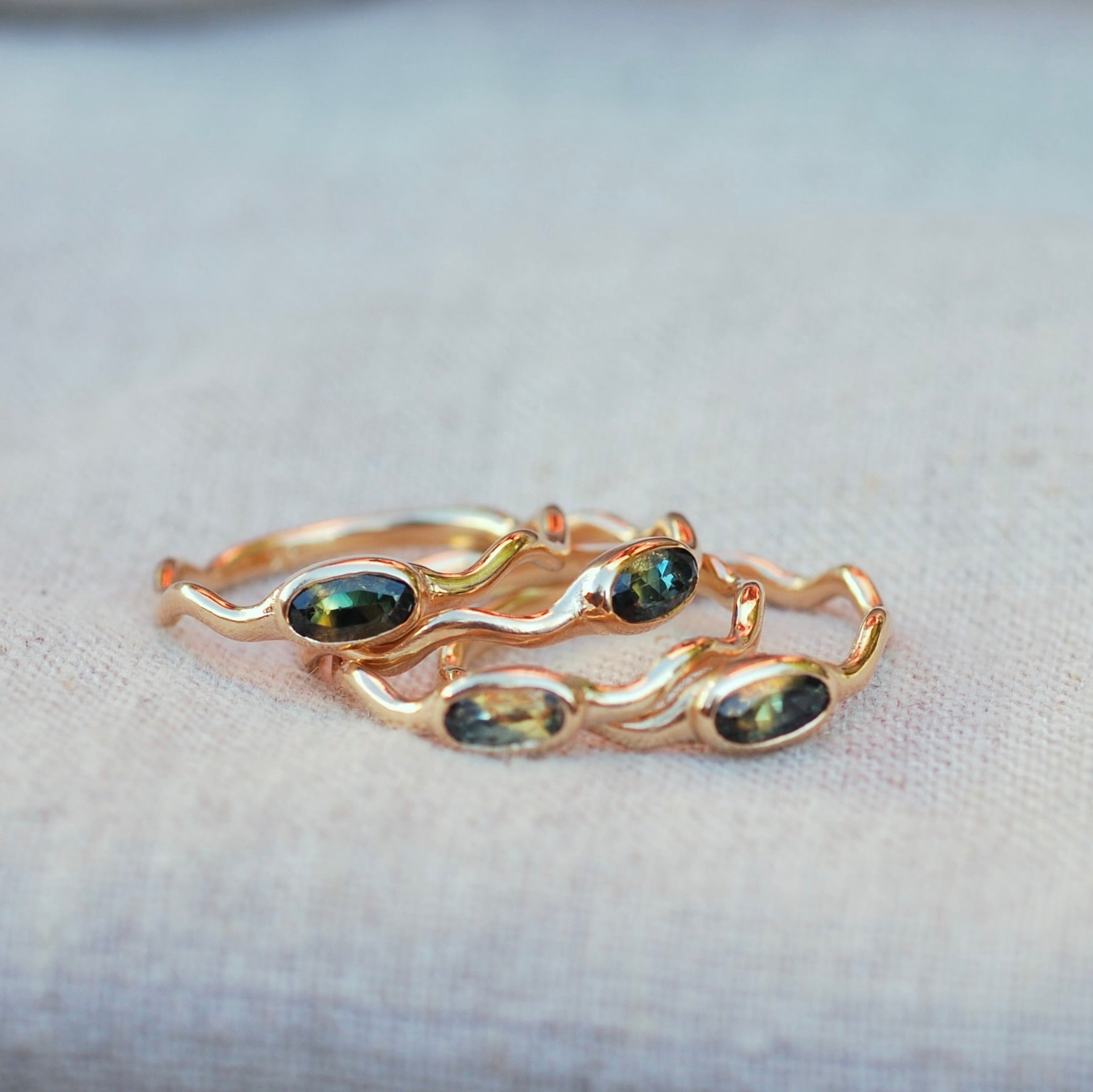 Gold rings with wavy bands and blue and yellow bi-color sapphires bezel set, laying on a beige fabric background