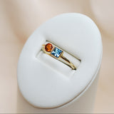 Gold ring with blue and orange gems on a white stand against a beige background