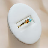 Gold ring with blue and orange gemstones on a white stand against a beige background