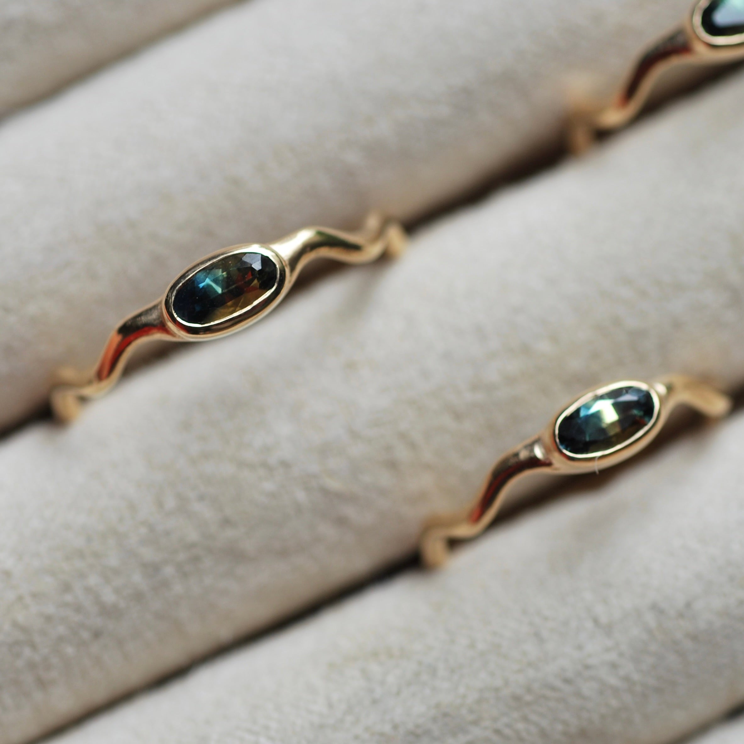 Gold rings with a horizontal oval bi-color sapphire and a wavy band in a beige ring tray