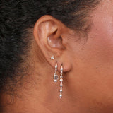 A person wearing two huggie hoops in silver, both with geometric charms