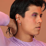 A person wearing silver huggie hoops with a geometric charm dangling from them and a silver thick ear cuff