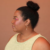 A person wearing gold colored earrings, a threader with a cube and sphere charm at the bottom and a huggie with the same charm