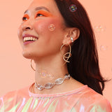 A woman wearing a pink top, bronze knot earrings, and a silver geometric necklace that resembles a spiral pattern