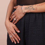 Hands and wrists wearing a cloud shaped bracelet, a squiggly bracelet, and multiple rings in gold and silver colors on a black background