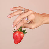 A hand holding a strawberry wearing a bronze signet with a purple amethyst teardrop stone and a mother of pearl circle stone