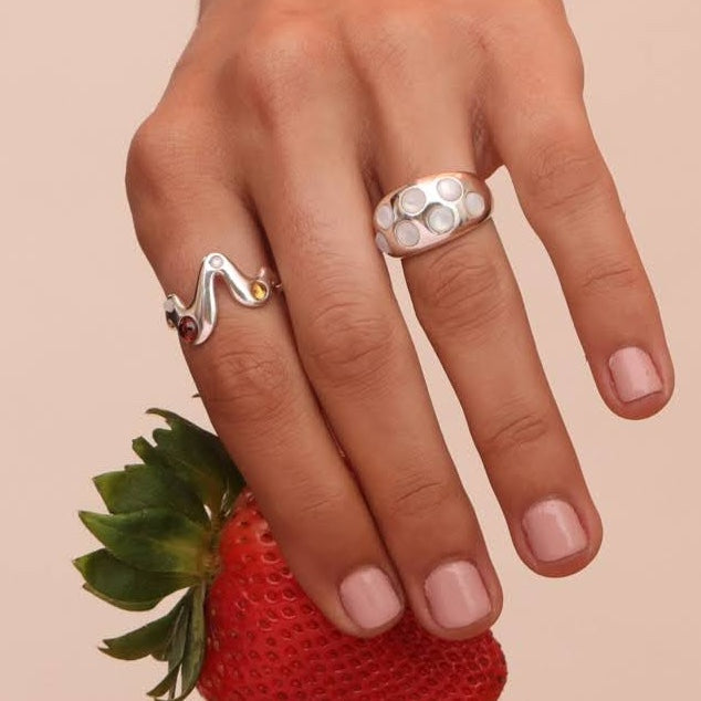 Close up of a hand wearing a silver drip ring with red, yellow, and white stones with a wide confetti ring with mother of pearl stones