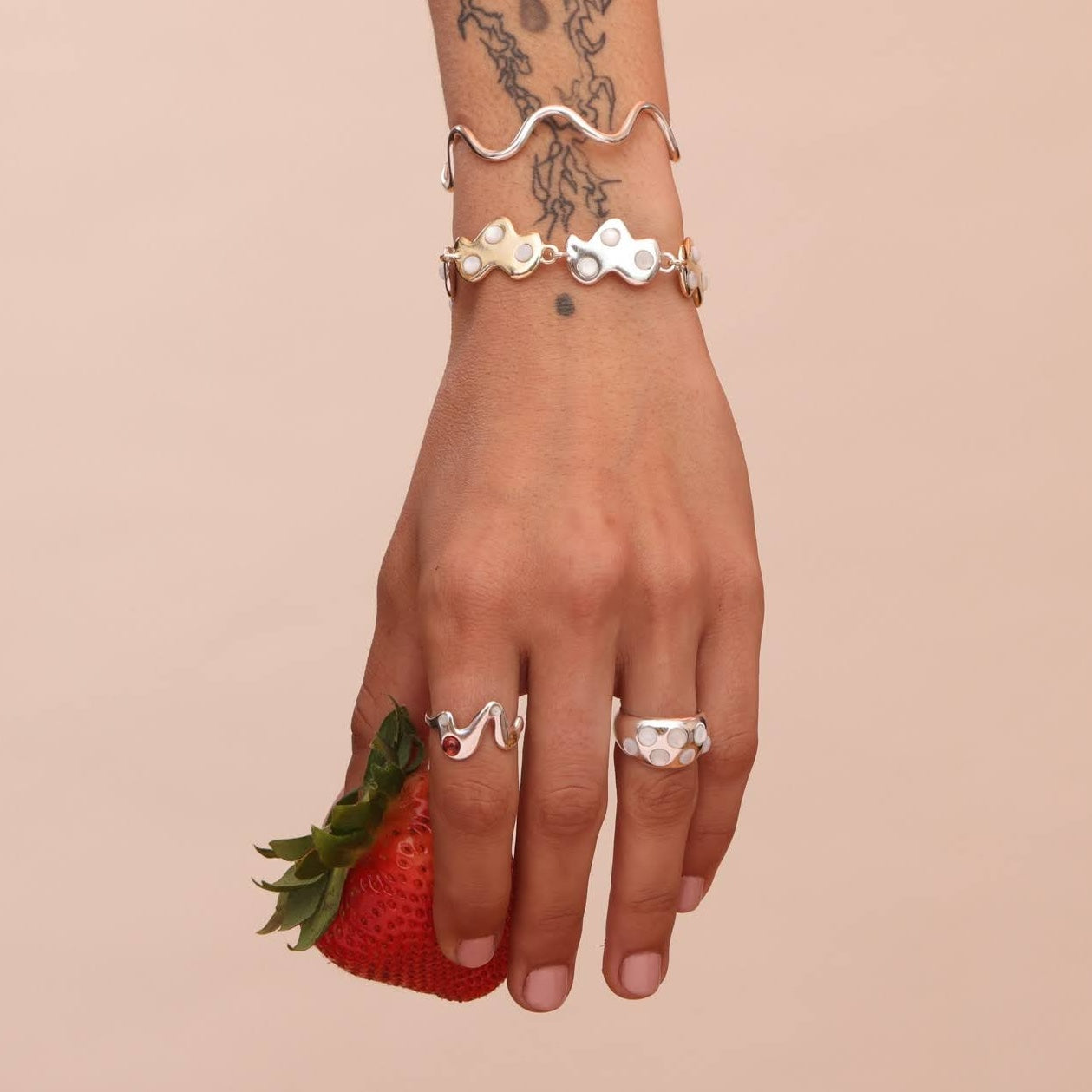 A person's hand displaying a silver ring with a wavy design, accompanied by other rings and a bracelet