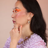 A photo of a person wearing a cloud shaped huggie earring featuring mother of pearl stones, and a bronze ear cuff