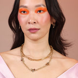A woman wearing a brass paperclip chain layered with another necklace that features cloud pendants and colored stones
