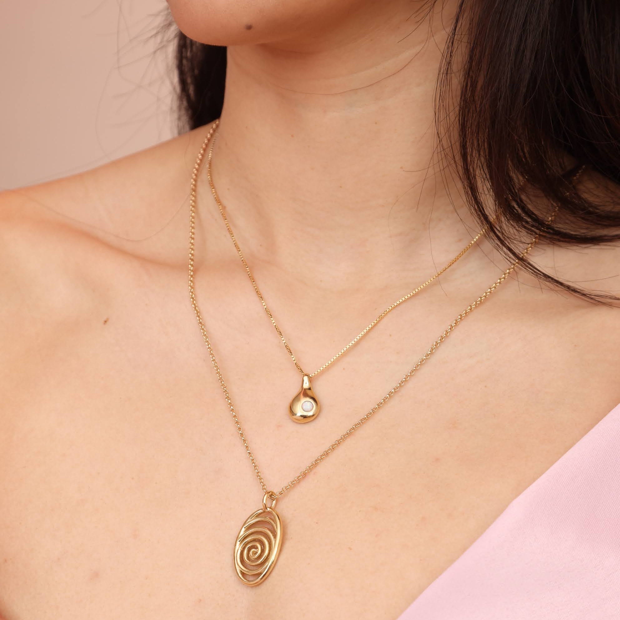 A person wearing a gold-colored necklace with a teardrop-shaped pendant with an opal stone stacked with another bronze necklace with a spiral pendant