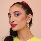 A woman wearing huggie earrings shaped like a cloud or artist palette in a gold bronze color with purple, teal, and opal stones