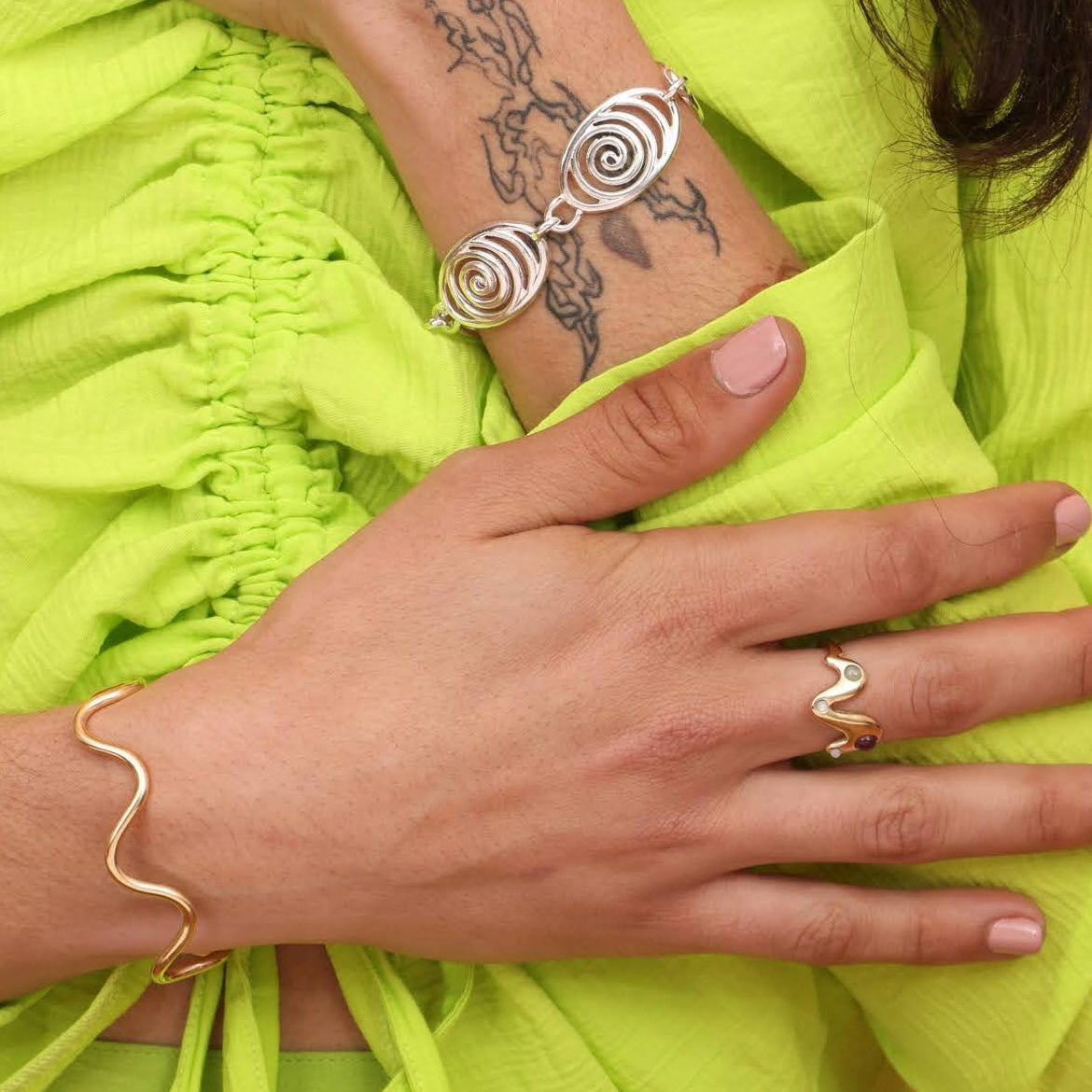 A person wearing a gold toned ring with colorful stones in it with a squiggly bracelet and a spiral bracelet