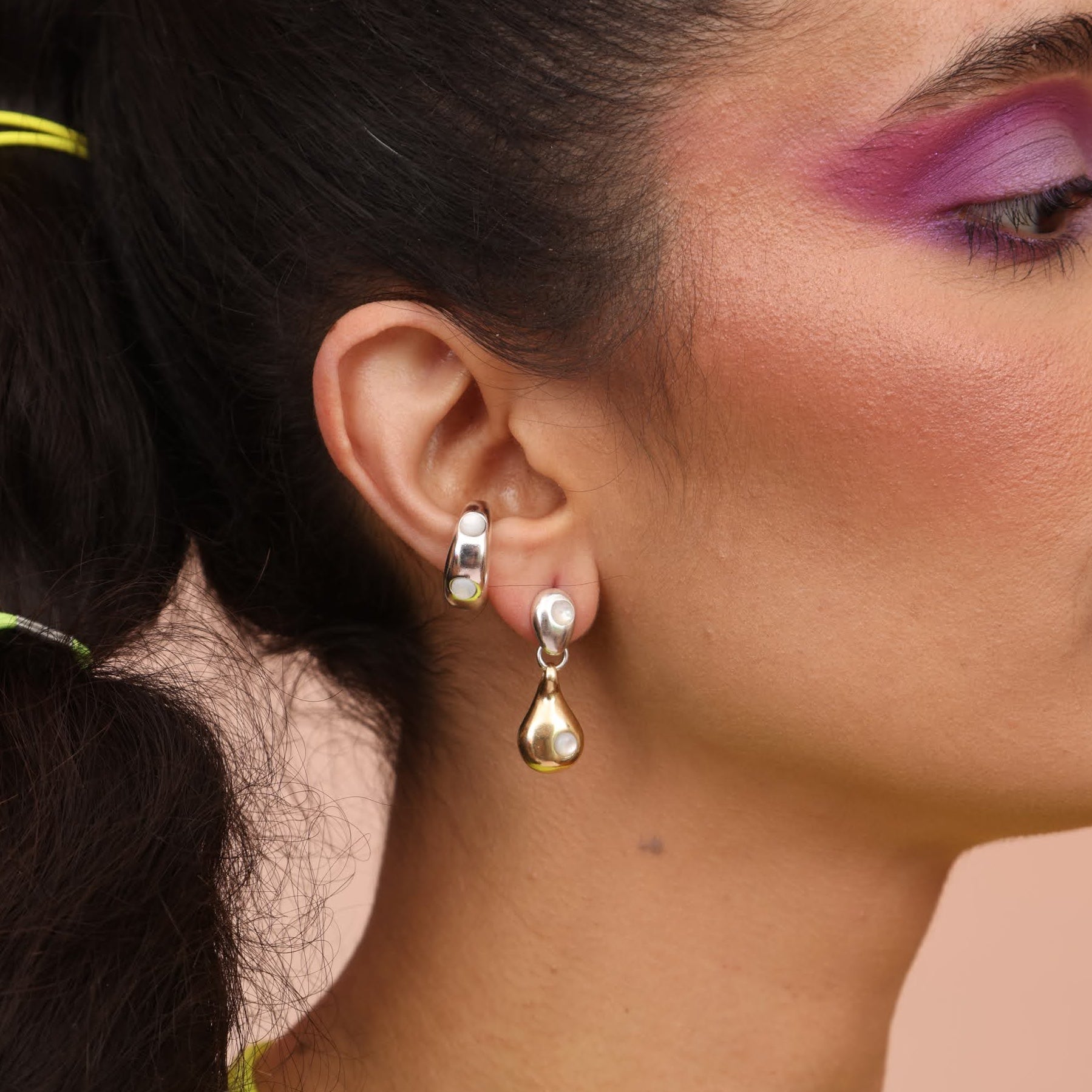 A close-up image of a woman's ear wearing a mixed metal drop-shaped dangle earring with mother of pearl and a silver ear cuff