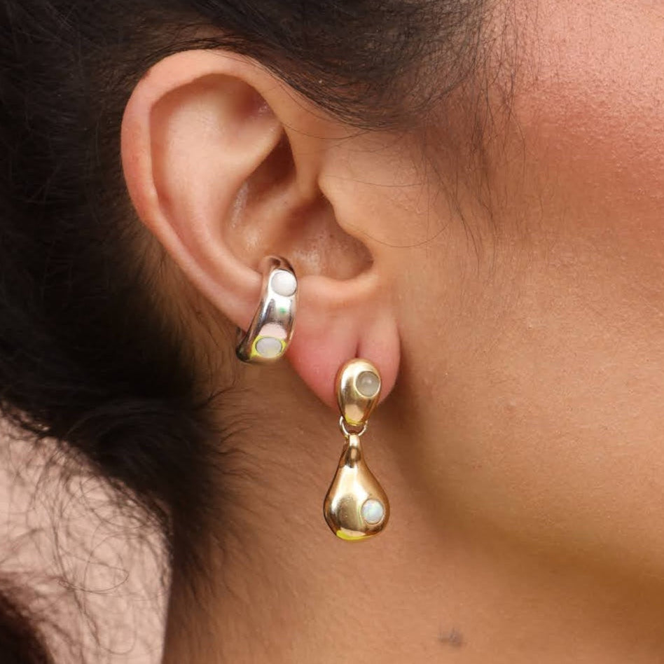 A close-up image of a woman's ear wearing a gold-colored drop-shaped dangle earring and a hoop ear cuff