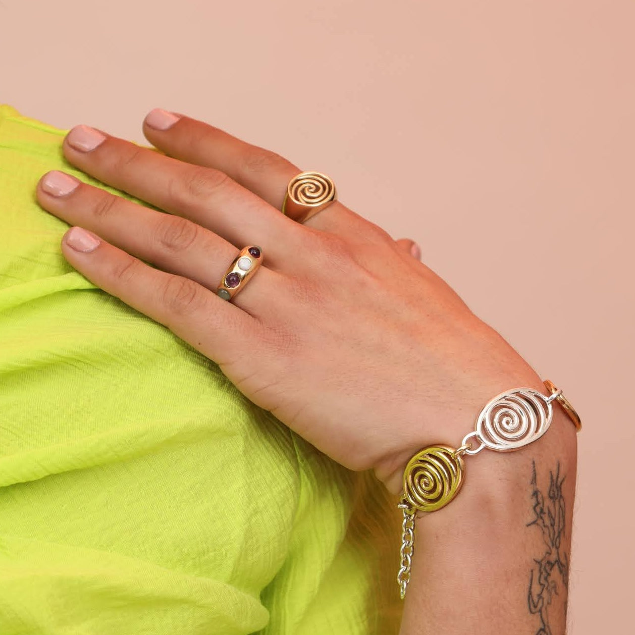 Person wearing an assortment of jewelry including a ring band with colorful cabochon stones, and a spiral signet and mixed metal spiral bracelet