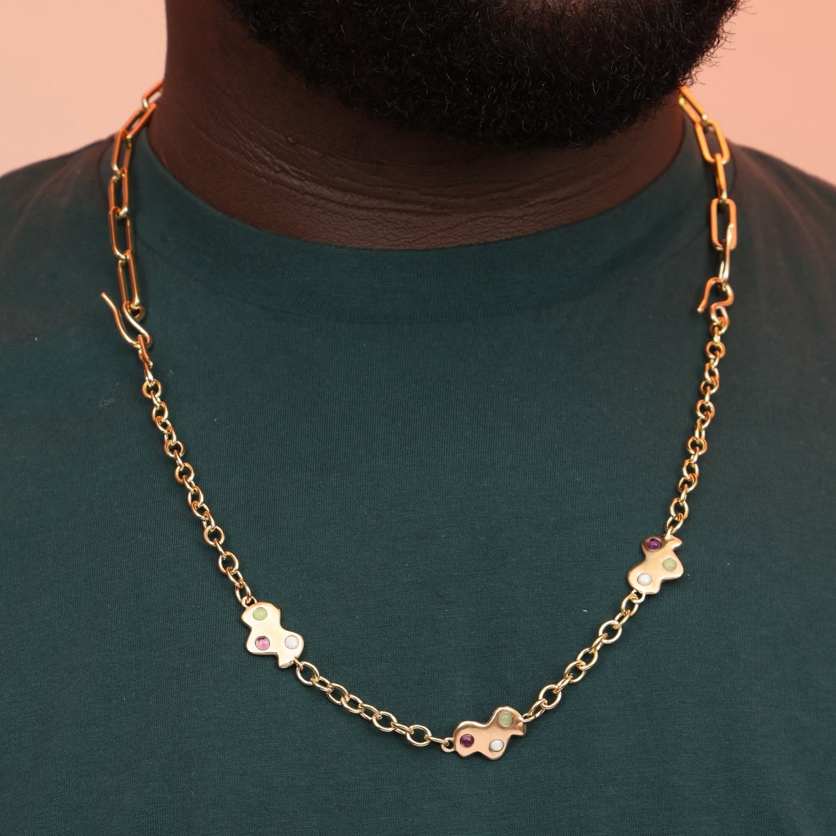 A person wearing a gold-colored necklace with three charms shaped like clouds, attached to a paperclip chain necklace, displayed against a green background.
