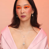A person wearing a spiral pendant necklace with a geometric design, stacked with a droplet necklace and displayed against a pink background.