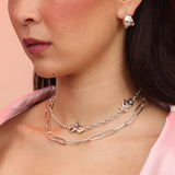 A person wearing a silver necklace with cloud charms and red, blue, and yellow stones layered with a paperclip chain necklace