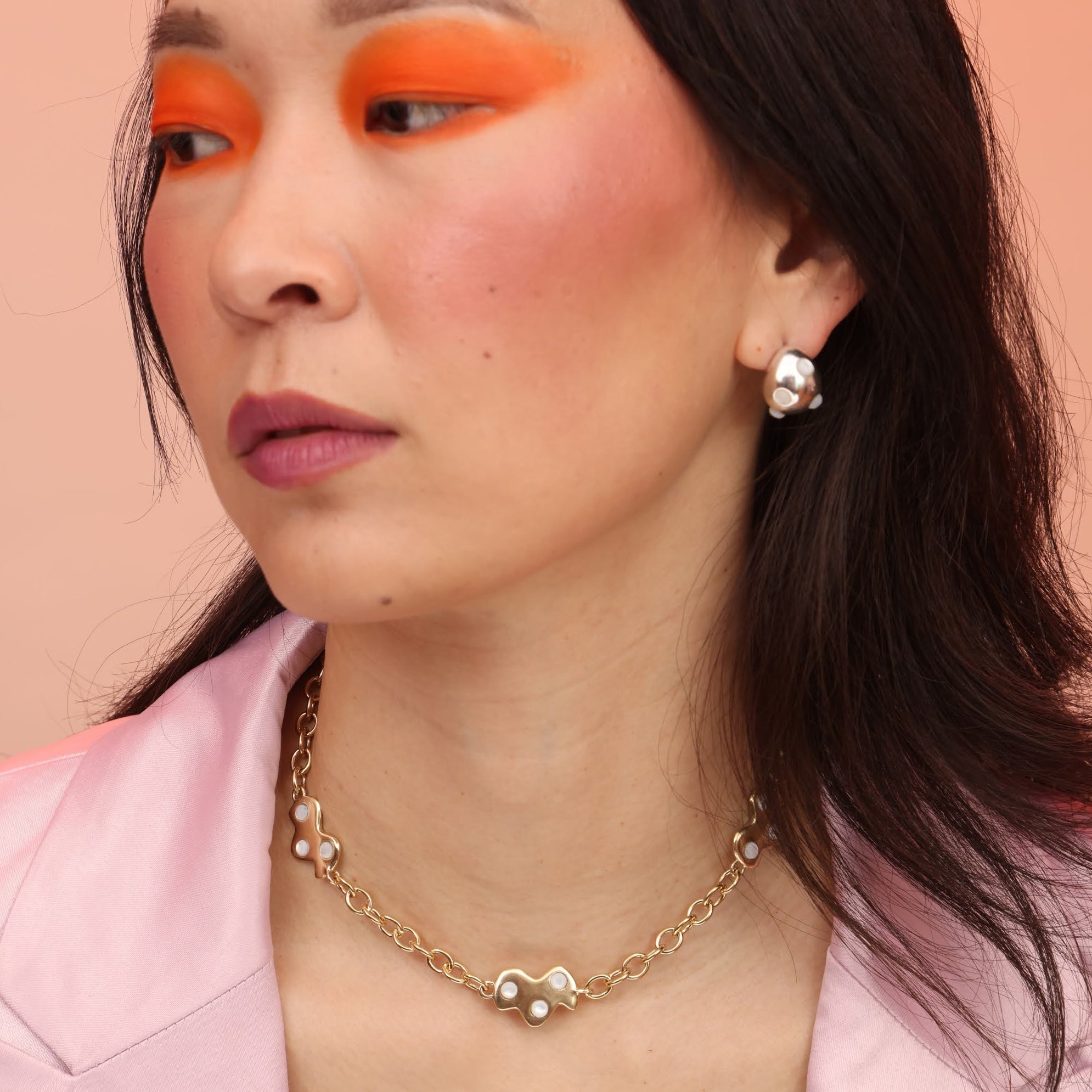 A person wearing a bronze gold necklace with cloud shaped charms and mother of pearl stones, as well as rounded hoops with pearl stones