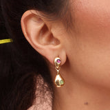 A close-up image of a woman's ear wearing a gold-colored drop-shaped dangle earring with amethyst and chrysoprase