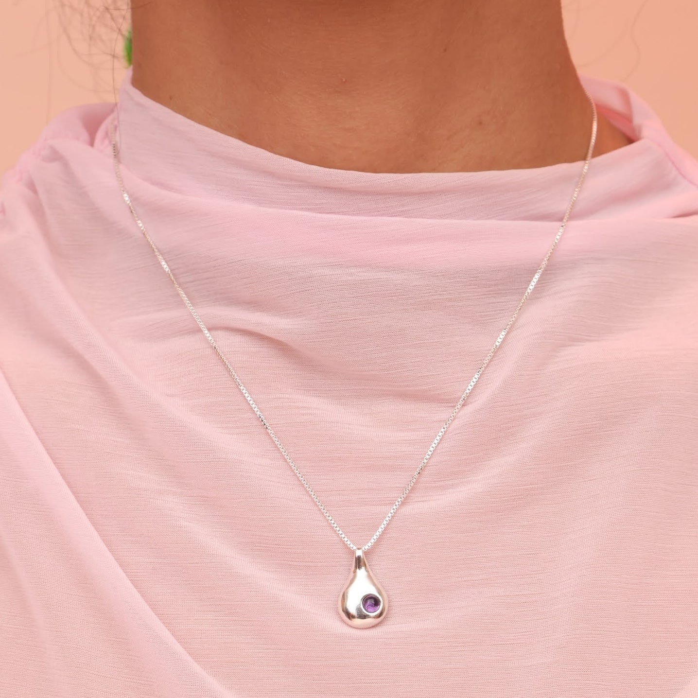 A person wearing a silver teardrop shaped necklace with a purple amethyst cabochon stone