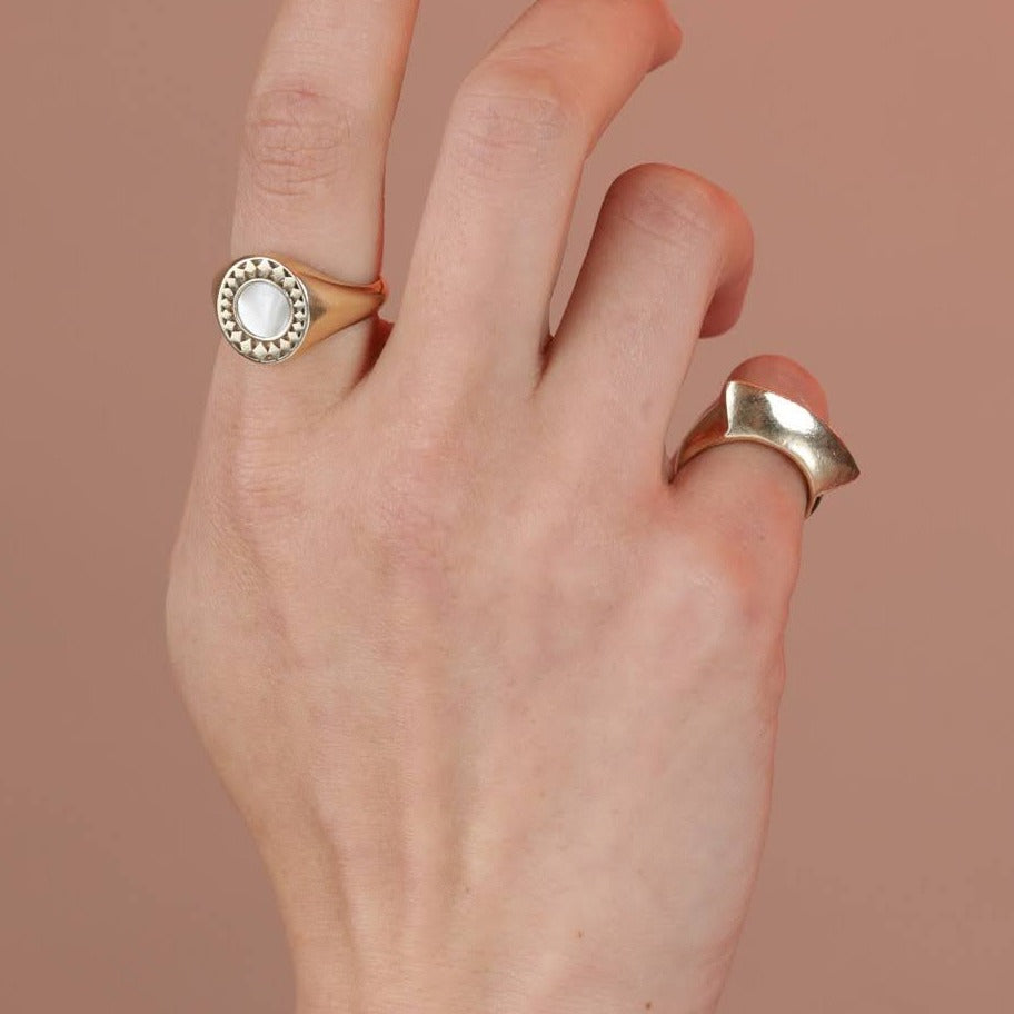 A close up of a hand wearing two gold toned bronze rings, one with a diamond border around a mother of pearl inlay, and one with fan like geometric shape
