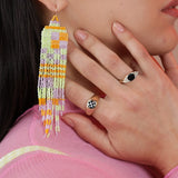 A person wearing colorful neon beaded earrings and gold colored bronze rings with checkerboard and black onyx inlay stones