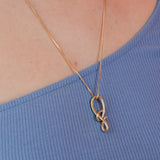 A gold-colored charm knot necklace on a 22-inch chain, displayed against the neckline of a blue ribbed garment.