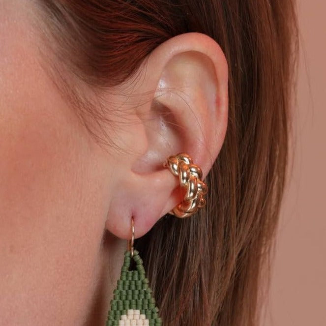 A person wearing a gold-colored braided ear cuff on their ear, with a green earring visible below.