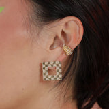 Close up shot of a person wearing gold colored bronze square checkerboard earrings and a ridged ear cuff