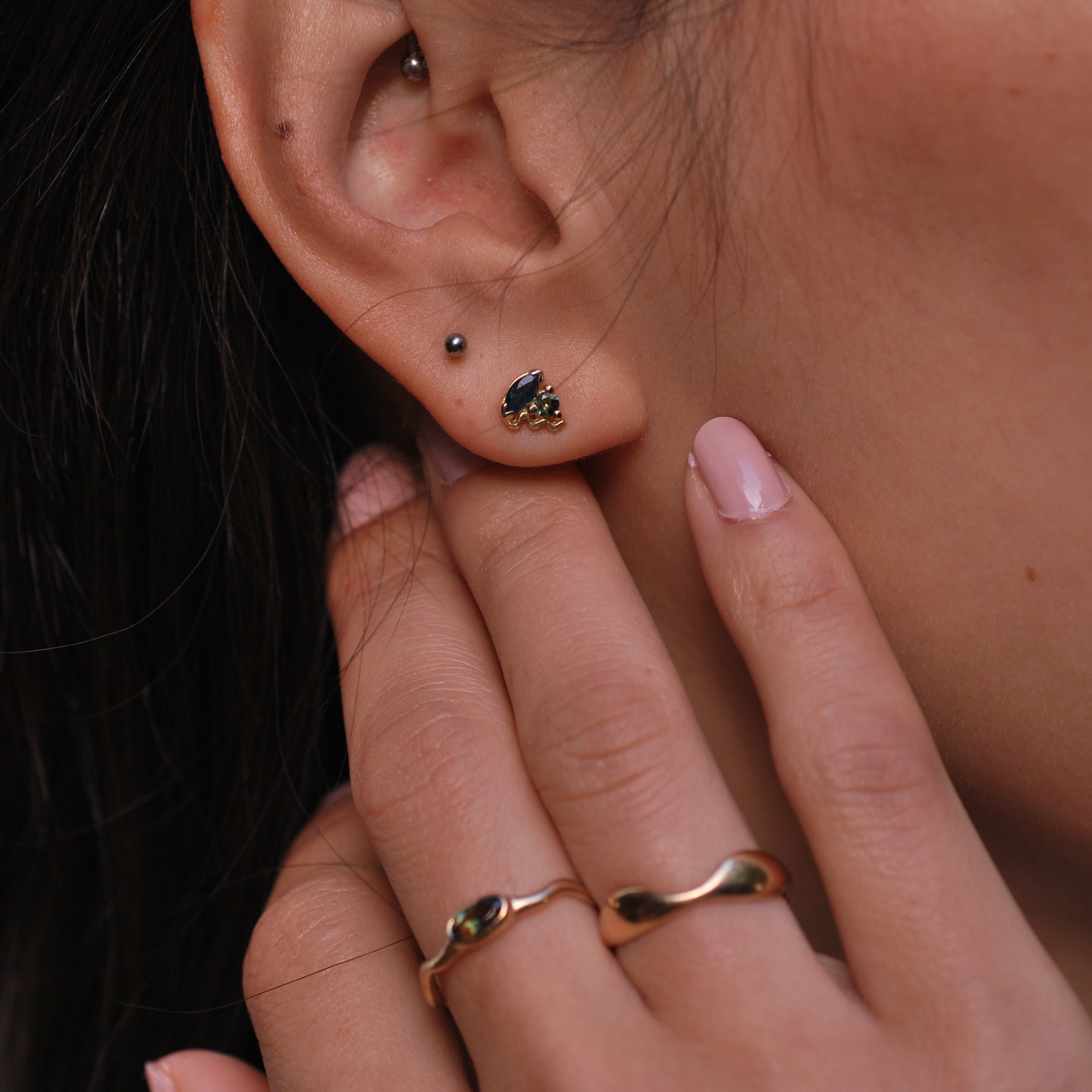 Close-up of an ear with a small earring with blue stones and a hand with rings on a blurred background