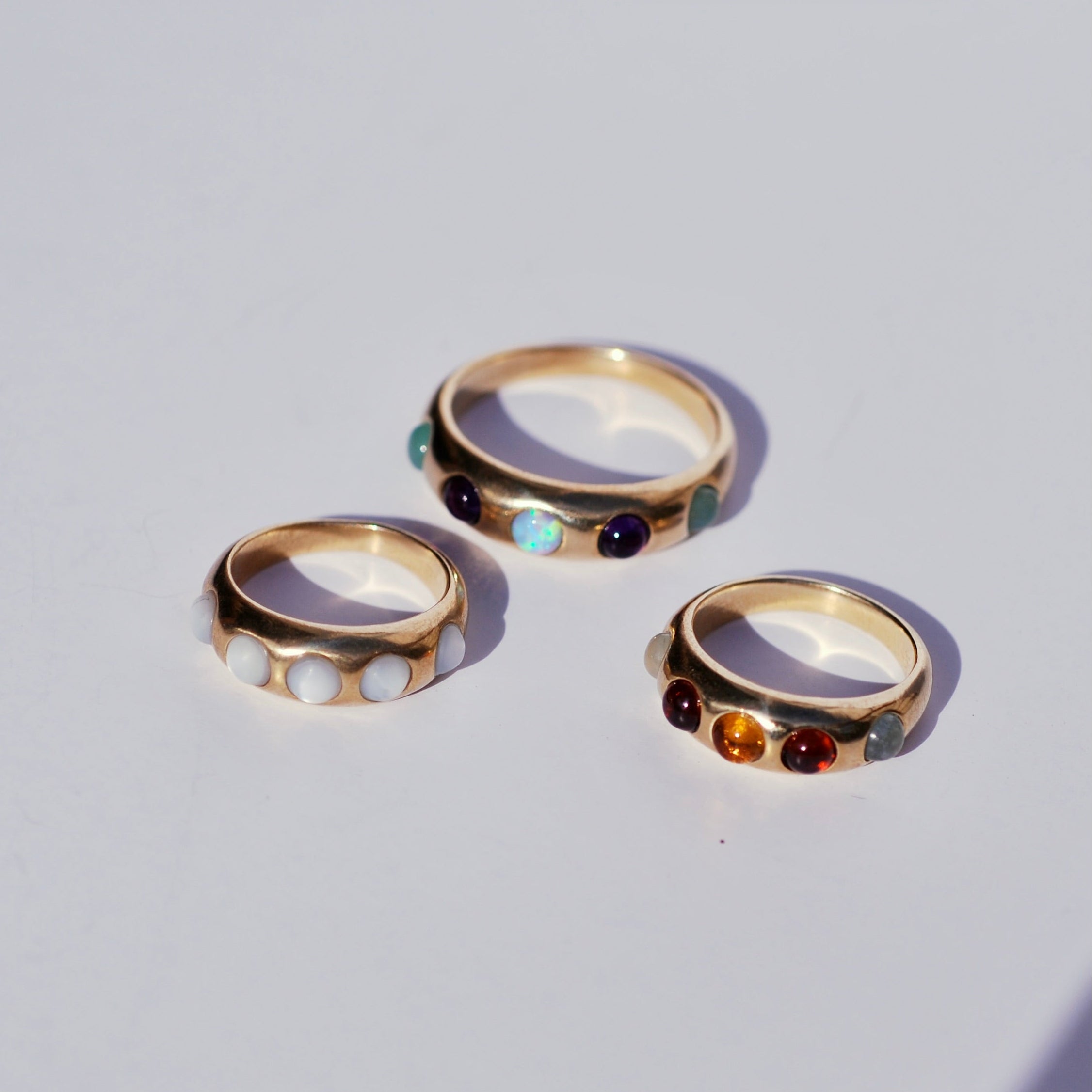 Three gold ring bands with colorful gemstones on a light gray background