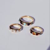 Three gold ring bands with colorful gemstones on a light gray background